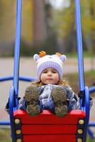 Baby girl in a funny hat swinging on the winter playground, perspective point of view. Looking at the camera photo