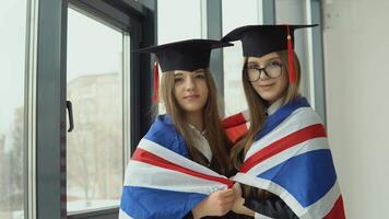 Two young happy graduate women stand shoulder to shoulder with a British flag on their shoulders video