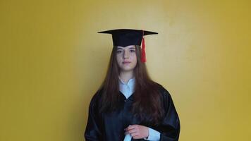 Caucasian femal student in a black gown and hat on a yellow solid background looks at the camera and smiles video