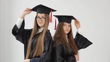 Two young happy graduate women stand shoulder to shoulder correct their hats with their hands. Close up view on white background video