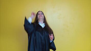 A university graduate stands in front of the camera holding a master's hat with a red tassel. Front shot of a young woman with long straight hair on a yellow background. Master of Divinity video