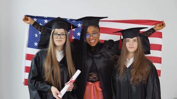 Tres joven contento graduado mujer de diferente nacionalidades estar juntos lado por lado y sostener diplomas y Estados Unidos bandera detrás a ellos. cerca arriba ver en blanco antecedentes video