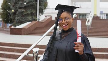 A young woman at the university at the master's mantle holds a diploma and looks at the camera. Portrait with the stairs in front of the central entrance to the university on background video