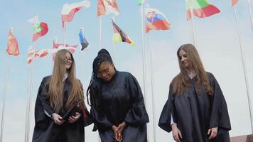 Tres joven mujer de diferente nacionalidades alegremente lanzar sombreros arriba en contra muchos banderas de diferente países revoloteando en el viento en el antecedentes. al aire libre ver video