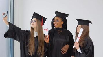 Tres joven contento estudiantes mujer de diferente nacionalidades estar juntos lado por lado y tomar un selfie con diplomas en su manos. cerca arriba ver en blanco antecedentes video