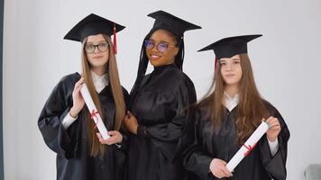 drei jung glücklich Absolvent Frauen von anders Nationalitäten Stand zusammen Seite durch Seite mit Diplome im ihr Hände. schließen oben Aussicht auf Weiß Hintergrund video