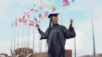 Young african american woman rejoices with a diploma of higher education in hand on the street against the background of flags of different countries in Europe and the world video