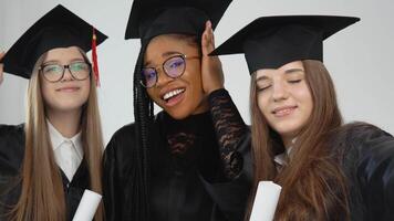 Tres joven contento graduado mujer de diferente nacionalidades estar juntos lado por lado con diplomas en su manos. cerca arriba ver en blanco antecedentes video