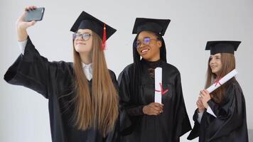 drie jong gelukkig afstuderen Dames van verschillend nationaliteiten staan samen en nemen een selfie met diploma's in hun handen. dichtbij omhoog visie Aan wit achtergrond video