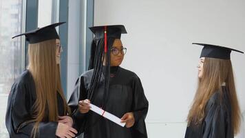 drie vrouw studenten van verschillend races met een diploma in hun handen. afgestudeerden in zwart Jurken en plein hoeden hebben een gesprek video