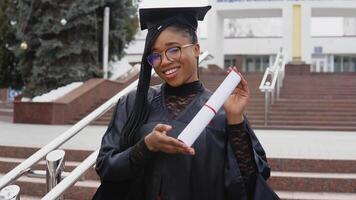 un joven mujer a el Universidad a el maestría manto sostiene un diploma mientras felizmente bailando y mira a el cámara. retrato en frente de el central Entrada a el Universidad en antecedentes video