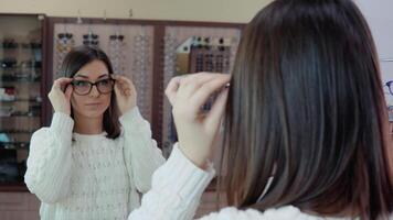 Jeune brunette avec bleu yeux dans une confortable blanc chandail en essayant sur des lunettes dans de face de une miroir dans un optique boutique video