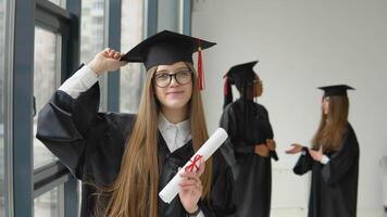 froh Absolvent mit einer Hand hält ein Diplom, und mit das andere Hand hält ein Hut auf das Hintergrund von zwei Studenten von anders Rennen. höher Bildung ist verfügbar zu Mittelklasse Frauen video