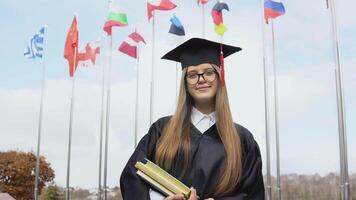 un Universidad graduado soportes en el antecedentes de banderas de el mundo participación libros en su manos. ver de el abierto espacio. nacional banderas en el azul cielo video