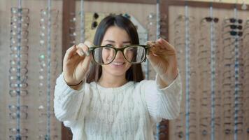 A young brunette with long hair in a white cozy sweater stands on the background of a shelf in an optics store and looks through glasses, holding them in outstretched arms video