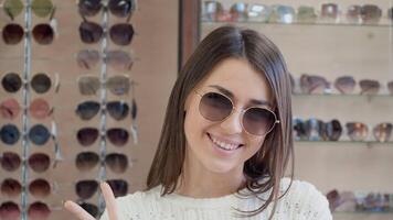 Cheerful young woman in sunglasses smiling on the background of a shelf with glasses in the store. video