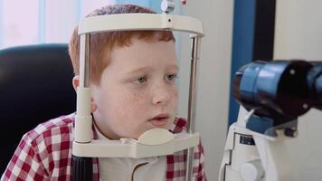 A red-haired caucasian boy with braces in a checkered red and white shirt in the ophthalmologist's office in the optics clinic undergoes a vision test using an autorefractometer video