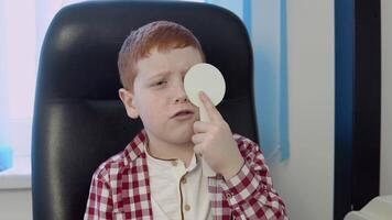 een roodharig jongen in een plaid rood en wit overhemd in de oogarts kantoor Bij de optiek kliniek antwoorden een dokter vraag gedurende een visie testen. video