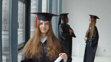 un sonriente graduado sostiene un diploma de mayor educación en su manos con otro hembra estudiantes en antecedentes. internacional estudiante intercambiar programa para socialmente vulnerable grupos video