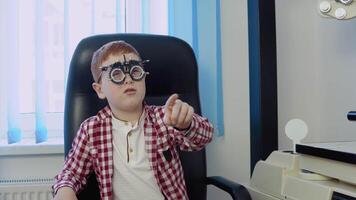 ein rothaarig Junge mit Hosenträger im ein kariert rot und Weiß Hemd im das Augenarzt Büro beim das Optik Klinik Antworten ein Arzt Frage während ein Vision prüfen. video