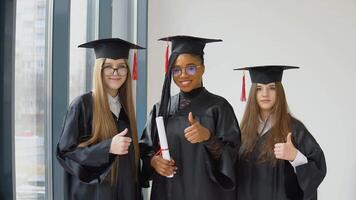 tre femmina studenti di diverso gare con un' diploma nel mano. laureati nel nero Vestiti e piazza cappelli Guarda a il telecamera con un' Sorridi. mille dollari per più alto formazione scolastica nel Europa e il unito stati video