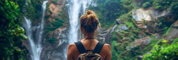 AI generated A young woman with a backpack standing in front of a powerful waterfall, experiencing the force of nature. photo