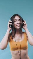 hermosa niña disfrutando música y poses para el leva. bonito joven mujer bailando y escuchando música con negro auriculares. amarillo ropa interior. azul antecedentes. vertical vídeo para teléfono. lento movimiento. video