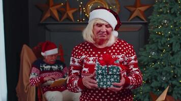 Mature grandmother in festive sweater presenting Christmas gift box, smiling, looking at camera video