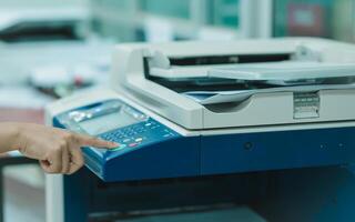 A employee is pointing at a printer, which is on and ready to print. The staff is likely preparing to print something, such as a document or a photo