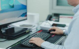 A engineer or programmer is typing on a computer keyboard in front of a monitor. He is wearing a blue shirt and a watch, coworking photo