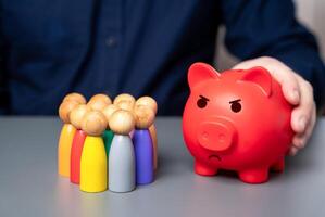 A businessman holds a red piggy bank next to a group of people figures. Lack of budget funds. Poor deposit conditions. Problems with pensions and savings systems. photo