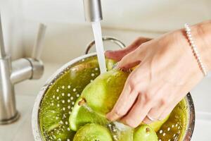 mujer en blanco camisa Lavado verde peras debajo claro agua en inoxidable acero cocina lavabo foto