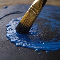 The image captures a close-up view of a paintbrush with golden hardware, applying vibrant blue paint onto a surface. The texture and glossiness of the wet paint are prominently displayed photo