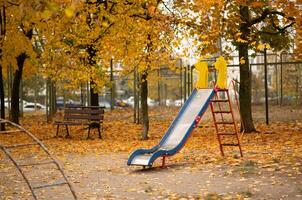 vacío patio de recreo con un hermosa naranja y rojo otoño antecedentes. antiguo niños diapositiva con arce hojas a otoño foto