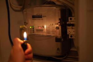 A man's hand with a lighter in complete darkness looks at the electric meter at home. Power outage, blackout concept. photo