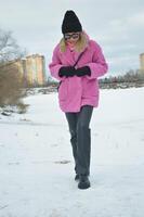 un de moda niña en un rosado piel Saco y un negro sombrero camina en el nieve en un ciudad parque. foto