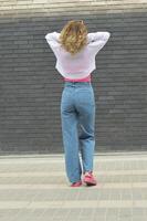 un rubia niña en pantalones y un blanco suéter soportes con su espalda a el cámara en contra un gris ladrillo pared. foto