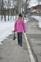 un de moda caucásico rubia niña en un brillante rosado invierno piel abrigo, un negro sombrero y pantalones camina a lo largo un asfalto la carretera en un invierno parque. foto