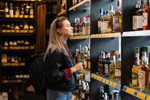 Ucrania. Jarkov octubre 2019 joven hermosa caucásico niña en un espíritu Tienda lo hace no Mira a un estante con botellas de whisky. foto