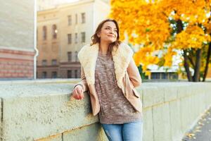 Beautiful caucasian brunette girl standing warm autumn day with background of trees with yellow foliage and a city. photo