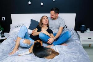 Young couple sitting on a bed in the bedroom, hugging. Near them is their dog photo