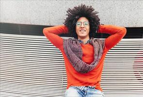 A handsome young Egyptian curly guy sitting on a bench dressed in a bright orange sweater and jeans. Happy people. A foreign student cupped his hands behind his head photo