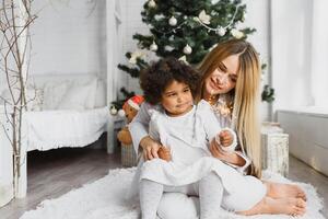 Merry Christmas and Happy Holidays Cheerful mom and her cute daughter at Christmas tree. Parent and little child having fun near Christmas tree indoors. Loving family with presents in room. photo