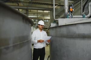 A young handsome employee of a modern factory in a white hard. In the background is a large shop for the production. photo