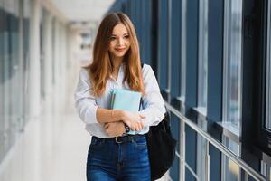 Positivity beautiful girl smiling at camera, standing on corridor with notes as backpack, going to lesson. Happy brunette female student studying in luxury university. photo