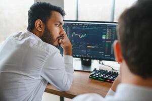 Side view of smart Indian crypto trader, investor, analyst broker, using laptop and smartphone analyzing digital cryptocurrency exchange, stock market charts, thinking of investing and funds risks. photo