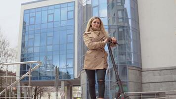 jung stilvoll blond Mädchen im ein Beige Graben Mantel und Jeans steht mit ein elektrisch Roller auf das Hintergrund von ein modern Hochhaus video