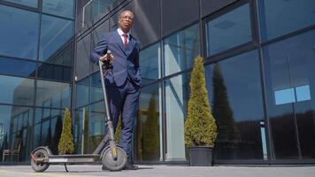 un africano americano en un clásico azul negocio traje soportes con un eléctrico scooter en el calle cerca un moderno espejo edificio con su mano en su bolsillo. fondo ver en lleno longitud video