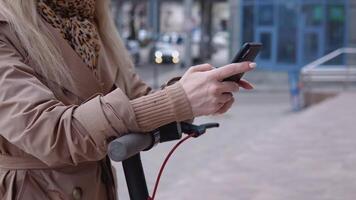 jung stilvoll blond Mädchen im ein Beige Graben Mantel und Jeans steht mit ein elektrisch Roller mit ein modern Hochhaus auf das Hintergrund und hält ein Zelle Telefon von ihr Tasche video