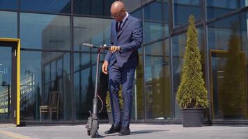 An African-American man in a classic blue business suit gets on an electric scooter and rides on the street near a modern mirror building. Bottom view in full length video